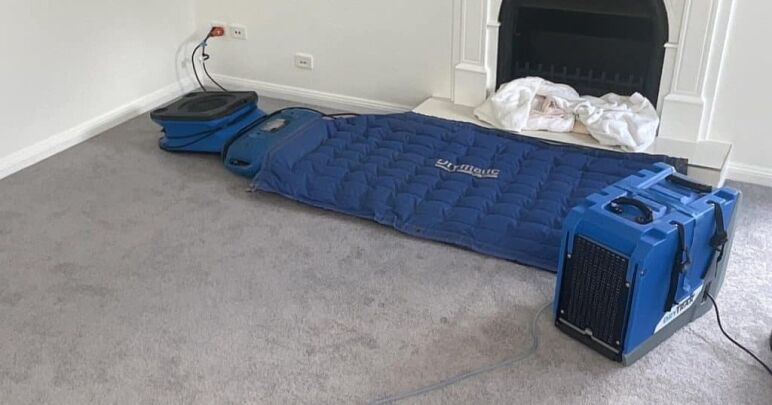 Blue Drymatic drying mat, air mover and dehumidifier set up on grey carpet beside a white fireplace